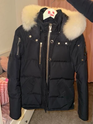 Svart dunjacka med pälskrage Moose Knuckles - Snygg svart dunjacka från Moose Knuckles med stor fluffig pälskrage i vitt. Jackan har flera dragkedjefickor, tryckknappar och en huva. Klassisk puffad design och logga på ärmen. Perfekt för kalla vinterdagar.