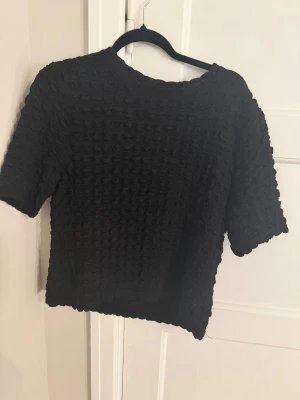 Svart strukturerad topp från Lindex - Svart topp från Lindex med kort ärm och coolt strukturerat tyg som ger en unik look. Storlek L men sitter mer som en S/m Modellen är rak och något croppad, perfekt till både jeans och kjol. Enkel att styla och passar till många olika outfits.