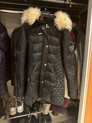  Rockandblue jacka  - Svart lång dunjacka från Rockandblue med snygg quiltad design och avtagbar huva med ljus päls. Jackan har flera dragkedjefickor, tryckknappar och Rockandblue-logga på ärmen. Perfekt för kalla vinterdagar. Använd en vinter 