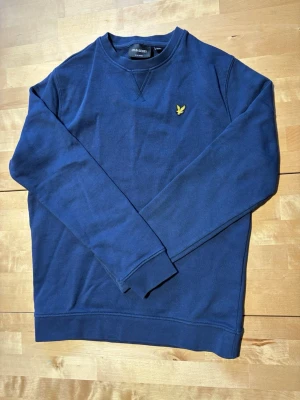 Mörkblå sweatshirt från Lyle & Scott - Mörkblå sweatshirt från Lyle & Scott med klassisk rund halsringning och gul logga på bröstet. Tröjan har långa ärmar, ribbade muddar och är tillverkad i mjukt bomullsmaterial. Perfekt för en avslappnad och stilren look.