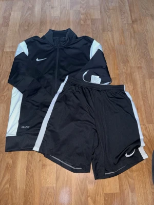 Svarta Nike träningsshorts och jacka - Säljer ett set med svarta Nike träningsshorts och matchande jacka med vita detaljer. Båda plaggen har Nike-loggan och är tillverkade i lätt, ventilerande Dri-FIT material som passar perfekt för träning eller sport.