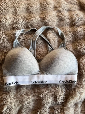 Grå sport-bh från Calvin Klein - Grå sport-bh från Calvin Klein med vadderade kupor och korsade axelband. Resårbandet nedtill har logotypen tryckt runtom. Mjuk och stretchig bomullsblandning för extra komfort. Vet inte helt säkert vilken storlek