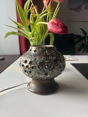 Keramisk elektrisk ljuslykta - Unik elektrisk ljuslykta i keramik med rund form och många små hål för ljusspridning. Färgen är en mix av brunt, blått och grönt med en glaserad yta. Lyktan har en bred fot och ansluts med sladd. Perfekt som mysig belysning i rummet.