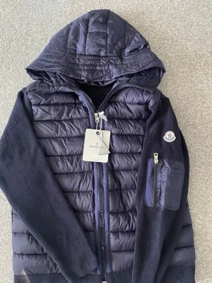  Moncler cardigan  - Snygg mörkblå dunjacka från Moncler med quiltad front och huva. Jackan har stickade ärmar och rygg, dragkedja framtill och en ficka med dragkedja på ärmen. Klassisk Moncler-logga på vänster ärm och innerfoder med serietidningsmotiv.