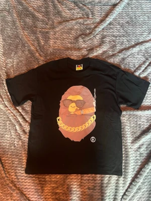 Svart BAPE t-shirt med tryck - Säljer en svart t-shirt från BAPE med ett stort tryck av en apa med solglasögon, guldkedja och cigarr på bröstet. Klassisk passform och korta ärmar. Ikonisk BAPE-logga på ärmen. Perfekt för dig som gillar streetwear och vill sticka ut.