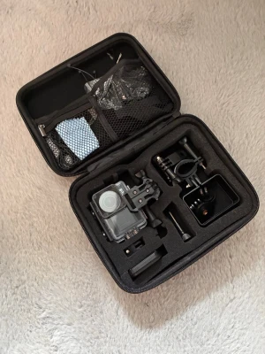 Raven actionkamera med tillbehör - Säljer en svart Raven actionkamera i robust plastmaterial, levereras i en hård skyddsväska med flera fack och tillbehör som fästen och laddare. Perfekt för dig som vill filma äventyr och sport. Allt är snyggt organiserat i väskan.