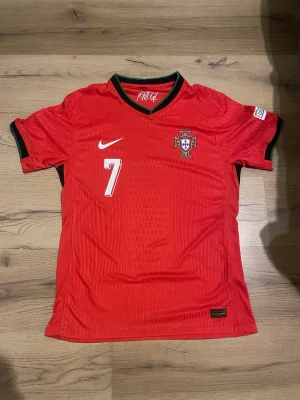 Portugal fotbollströja Ronaldo #7 Nike - Röd Portugal fotbollströja från Nike med nummer 7 och Ronaldo på ryggen. Tröjan har gröna detaljer vid krage och ärmslut, samt landslagets emblem på bröstet. Tillverkad i lätt och ventilerande material, perfekt för träning eller match. Slim fit 