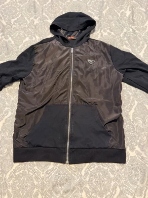 Svart hoodie från Prada med dragkedja - Snygg svart hoodie från Prada med dragkedja framtill och klassisk huva. Framsidan är i glansigt material med Pradas ikoniska logga på bröstet. Stora fickor framtill och ribbade muddar. Perfekt för dig som gillar exklusiva streetwear-plagg.