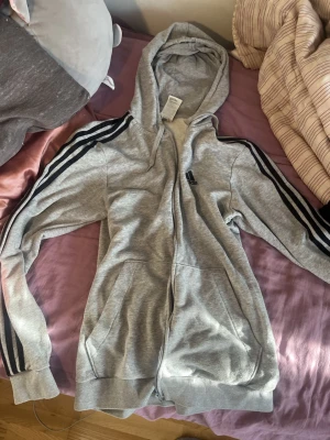 Grå Adidas hoodie med dragkedja - Snygg grå hoodie från Adidas med dragkedja framtill och klassiska svarta ränder längs ärmarna. Adidas-logga broderad på bröstet. Utrustad med huva och två fickor framtill. Perfekt för en sportig och avslappnad stil.