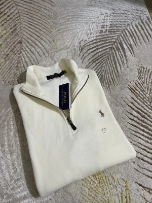 Vit halvzip från Polo Ralph Lauren - Snygg vit tröja från Polo Ralph Lauren med halv dragkedja och broderad logga på bröstet. Tröjan har ribbad struktur och hög krage, perfekt för en clean och stilren look. Passar till många olika outfits och är lätt att matcha.