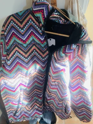 Färgglad zigzag mönstrad pufferjacka - Säljer en riktigt färgstark pufferjacka med zigzag-mönster i lila, rosa, blått, svart och turkos. Jackan har dragkedja framtill, två fickor och en hög krage. Perfekt för dig som vill sticka ut i vinter och gillar unika plagg med mycket attityd. Stor i storlek