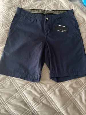 Marinblå shorts från Sail Racing - Snygga marinblå shorts från Sail Racing med klassisk passform och diskret logga bak. Tillverkade i ett lätt och slitstarkt material, perfekta för sommardagar. Två sidofickor och en bakficka ger praktisk förvaring. Stängs med knapp och dragkedja.