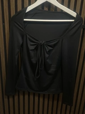Svart topp med knyt framtill - Trendig svart långärmad topp med djup urringning och knytband framtill. Sitter snyggt figurnära och passar perfekt till jeans eller kjol. Tillverkad i mjukt, stretchigt material som känns bekvämt hela dagen.