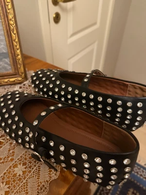 Svarta ballerinaskor med nitar -  Köpta för 5000. Snygga svarta ballerinaskor med rund tå och rem över foten. Skorna är täckta med silvriga nitar som ger en cool och edgy look. Tillverkade i konstläder med brunt innerfoder. Perfekta för dig som vill sticka ut med din stil.