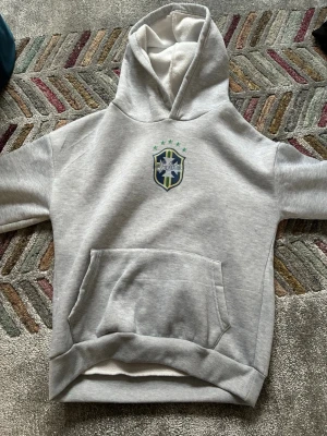 Grå hoodie med Saint-logga - Snygg grå hoodie från Saint med broderad logga på bröstet i blått, grönt och gult. Hoodien har en klassisk känguruficka framtill och en stor huva. Perfekt för en avslappnad och sportig stil.