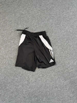 Svarta Adidas träningsshorts - Snygga svarta träningsshorts från Adidas med vita detaljer och klassiska ränder på sidan. Shortsen har elastisk midja med snörning och är tillverkade i ett lätt och svalt polyestermaterial, perfekta för sport och aktivitet.