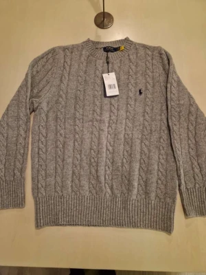 Grå kabelstickad tröja Polo Ralph Lauren - Kabelstickad grå tröja från Polo Ralph Lauren med rund halsringning och broderad marinblå logga på bröstet. Tröjan har ribbade muddar och nederkant, perfekt för lager-på-lager-stil. Klassisk och tidlös design.