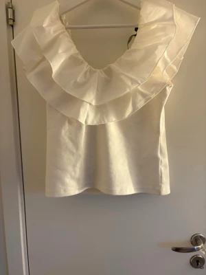 Vit volangtopp från Vero Moda - Supersnygg vit topp från Vero Moda med stora volanger runt axlarna och urringning. Toppen är ärmlös och har en croppad passform som ger en trendig look. Perfekt att styla med jeans eller kjol för en cool outfit.