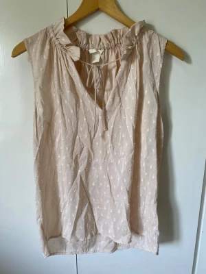 Ljusrosa ärmlös blus med volang - Söt ljusrosa ärmlös blus med små vita broderade prickar, volangdetalj vid halsringningen och knytband framtill. Blusen har en avslappnad passform och är tillverkad i ett lätt, luftigt tyg som passar perfekt till sommaren.