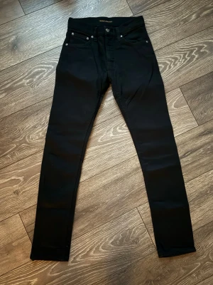 Nudie grim tim - Säljer ett par svarta nudie grim Tim jeans med klassisk femficksdesign och smal passform. Jeansen har hög midja och är tillverkade i stretchigt material för extra komfort. Perfekta för dig som gillar en clean och stilren look.