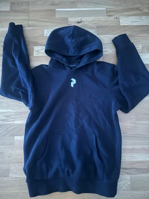 Svart hoodie från peak performance  - Svart hoodie med stor huva och magficka. På bröstet och ryggen finns ett stilrent grafiskt tryck i ljusblått. Hoodien har ribbade muddar vid ärmar och nederkant. Perfekt för en chill och avslappnad stil. Materialet känns mjukt och bekvämt, troligen bomull eller bomullsblandning.