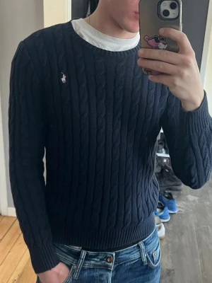 Mörkblå kabelstickad tröja Polo Ralph Lauren - Kabelstickad mörkblå tröja från Polo Ralph Lauren med rund halsringning och den klassiska broderade logotypen på bröstet. Tröjan har långa ärmar och ribbade muddar vid ärmslut och nederkant. Perfekt för en stilren och avslappnad look. Obs de är en dammodell men funkar för båda o sitter som xs herr