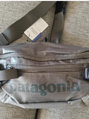 Helt ny grå magväska / axelremsväska från Patagonia - Snygg grå magväska som kan användas som axelremsväska från Patagonia med justerbart band och flera dragkedjefack samt sidofack. Tillverkad i slitstark nylon med stor logga framtill. Perfekt för att bära det viktigaste och enkel att matcha med olika outfits. 5 liter. 