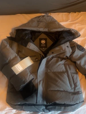 Canada goose Macmillian Stl S !Pris kan diskuteras! - Snygg grå dunjacka från Canada Goose med huva och svarta detaljer. Jackan har dragkedja, stora fickor och klassisk patch på ärmen. Perfekt för kalla vinterdagar och riktigt varm tack vare dunfyllningen. Pris kan diskuteras