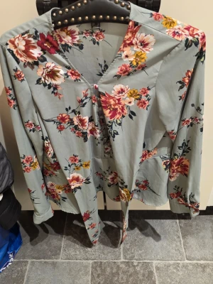 Grön blommig blus med knytband - Snygg blus i ljusgrönt med färgglatt blommönster i rött, rosa och gult. Blusen har v-ringning, långa ärmar och knytband i midjan för en feminin siluett. Tillverkad i ett lätt och mjukt tyg som ger en skön känsla.