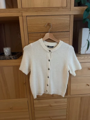 Vit kortärmad kofta med knappar - Söt vit kofta med korta ärmar och rund hals. Koftan har ribbade muddar och knäpps framtill med silvriga knappar. Perfekt att slänga över en topp för en clean och stilren look. Materialet ser mjukt och lite fluffigt ut.