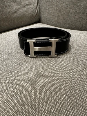 H-bälte - Snyggt svart bälte i läder med kontrastsömmar och ett stort, silverfärgat H-format metallspänne. Perfekt accessoar för att lyfta din outfit med en stilren och exklusiv touch. (Tillkommer även verktyg för att göra egna hal)