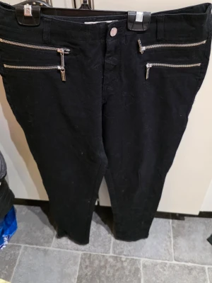 Svarta jeans från Michael Kors - Snygga svarta jeans från Michael Kors med fyra silverfärgade dragkedjor framtill som ger en cool och modern look. Jeansen har smal passform och är perfekta för dig som gillar en stilren men edgy stil. Materialet känns mjukt och stretchigt.