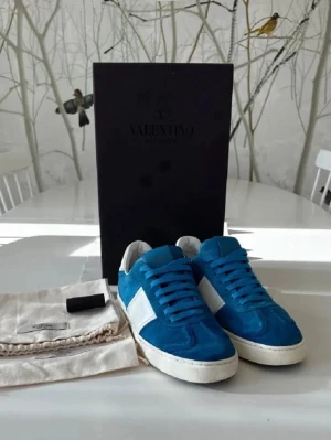 Blåa Valentino Flycrews sneakers med vit detalj - Snygga blåa sneakers från Valentino med vita detaljer på sidorna och vit sula. Skorna har klassisk låg modell och är tillverkade i mocka och läder. Perfekta för dig som vill sticka ut med stilrena och trendiga skor.