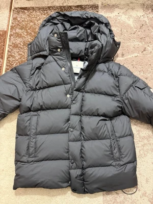 Svart dunjacka från Moncler - Snygg svart dunjacka från Moncler med stor huva och klassisk Moncler-logga på ärmen. Jackan har dragkedja och tryckknappar framtill samt två fickor med dragkedja. Puffig modell som håller dig varm under kalla dagar.