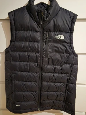 Svart dunväst från The North Face - Snygg svart dunväst från The North Face med quiltad design och hög krage. Västen har dragkedja framtill, bröstficka med dragkedja och logga på bröstet. Perfekt för lager-på-lager och håller dig varm med 550-fill dun.