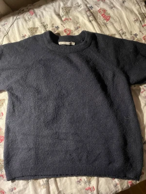 Mörkblå stickad tröja med rund hals  - Mysig mörkblå stickad tröja med rund halsringning och kortare ärmar. Tröjan har en enkel och stilren design, perfekt att matcha med jeans eller kjol. Materialet känns mjukt och värmande, och passformen är avslappnad. Den är kortärmad 