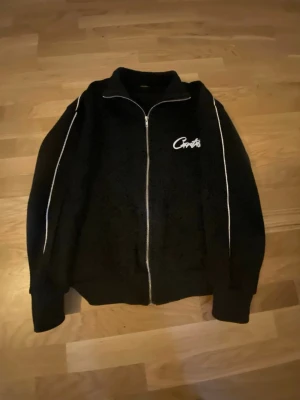 Svart ziptröja från Corteiz  - Svart ziptröja från Corteiz med vit logga på bröstet och vita ränder längs ärmarna. Tröjan har hög krage, hel dragkedja och ribbade muddar. Perfekt för en sportig och avslappnad stil.