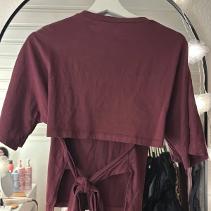 Vinröd croppad topp från Nelly - Säljer en vinröd croppad topp från Nelly med korta ärmar och rund hals. Toppen har en snygg öppen rygg med knyt-detalj som ger en cool vibe. Materialet är mjuk bomull och färgen är djup vinröd. Perfekt för dig som gillar trendiga detaljer!