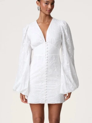 Vit kort klänning från Adoore - Supersöt vit kort klänning från Adoore med v-ringning och långa puffärmar. Klänningen har diskret blommigt mönster i tyget och knappar framtill. Perfekt för student eller skolavslutning