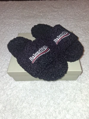 Svarta fluffiga tofflor från Balenciaga - Mysiga svarta tofflor från Balenciaga med broderad logga i vitt och rött framtill. Tillverkade i ett fluffigt, teddy-liknande material som ger en lyxig och trendig känsla. Perfekta för att chilla hemma med stil.