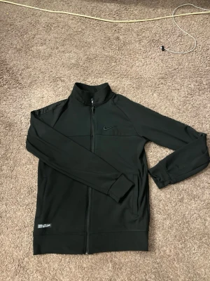 Svart ziptröja från Nike Storlek S - Svart ziptröja från Nike med diskret logga på bröstet och dragkedja hela vägen. Tröjan har hög krage, långa ärmar och ribbade muddar. Perfekt för en sportig och clean look. Priset kan diskuteras