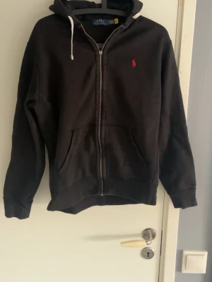 Svart hoodie från Polo Ralph Lauren - Svart hoodie med dragkedja från Polo Ralph Lauren. Klassisk design med huva och vita snören, två fickor framtill och rött broderat logotyp på bröstet. Mjuk bomullskänsla och perfekt för lager-på-lager-stil. Dragkädjan är i sönder så har sänkt priset mycket på grund av de.