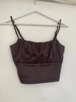 Brun croppad linnetopp med smala band - Snygg brun croppad linnetopp med smala justerbara axelband. Toppen har en enkel design med lätt rynkad detalj upptill och är perfekt för varma dagar eller att ha som fest topp. Tillverkad i mjukt material. Ifrån Hm.