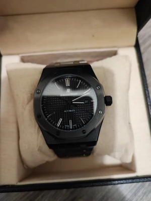 Svart Audemars Piguet Royal Oak klocka - Snygg svart Audemars Piguet Royal Oak automatisk klocka med ikonisk åttakantig boett och svart länkarmband. Urtavlan har ett rutigt mönster, datumvisning och silverfärgade index. Perfekt för dig som gillar exklusiv och stilren design.