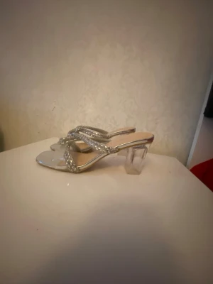 Silvriga pumps med klack och strass - Säljer ett par silvriga pumps från CULTURE med fyrkantig tå, transparent blockklack och glittriga remmar dekorerade med strass. Skorna har öppen häl och en modern, trendig look som sticker ut.
