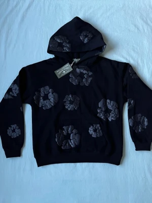 Svart hoodie från Denim tears - Säljer en svart hoodie från Denim tears med unika, upphöjda blommönster i mörkgrått över hela tröjan. Hoodien har en klassisk huva och en stor magficka framtill. Materialet känns mjukt och skönt, perfekt för en avslappnad streetwear-look.