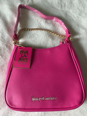 Juicy Couture rosa handväska - En helt ny handväska från Juicy Couture i starkt rosa färg. Den är aldrig använd och har även prislapp kvar. Väskan har guldfärgad kedjedetalj på axelremmen och logotyp framtill. Tillverkad i slätt konstläder och har en klassisk, rektangulär form. Perfekt för dig som vill sticka ut med en färgglad accessoar.