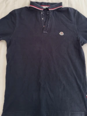 Marinblå pikétröja från Moncler - Snygg marinblå pikétröja från Moncler med klassisk krage och knappar framtill. Tröjan har Moncler-logga på bröstet och kragen har röda och vita detaljer. Perfekt till en avslappnad men stilren look.