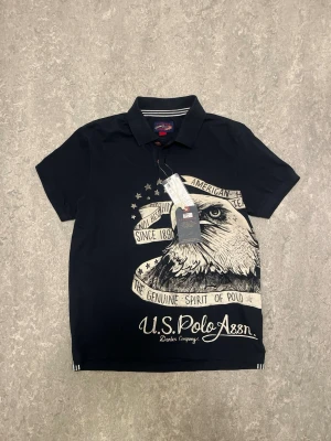 U.S. Polo Assn. Pike/Polo - Marinblå pikétröja från U.S. Polo Assn. med stor örn- och textprint framtill. Klassisk krage och korta ärmar. Snygg kontrast mellan Marinblå och vitt i trycket. Perfekt för dig som vill sticka ut med en cool och sportig look.