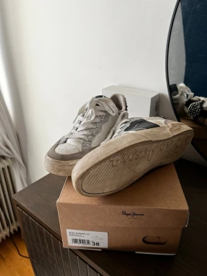 Vita och grå sneakers från Pepe Jeans - Snygga sneakers från Pepe Jeans i vitt och glittriga med svarta detaljer på sidan. Tyvärr för stora så inte använt många gånger, modellen är som den ser ut och ska vara ”smutsig” på sulan